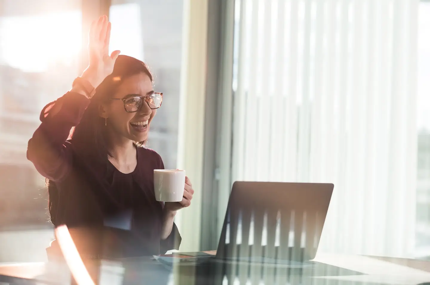 Smilende person med på jobb med laptop og kaffekopp løfter hånden for å hilse en kollega velkommen.